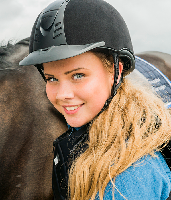 En ung jente med ridehjelm og blond parykk smiler mot kamera