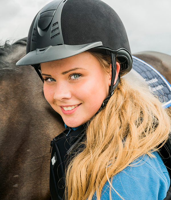 Tiril med ridehjelm og blond parykk står foran en hest