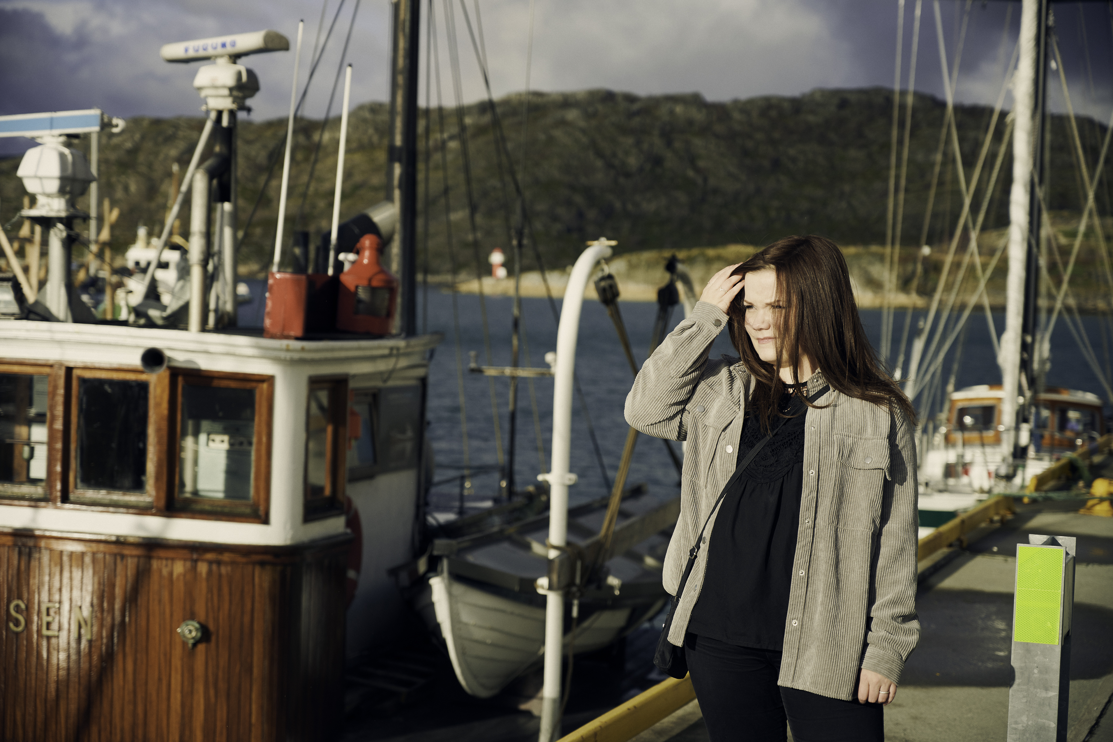 En ung kvinne med langt rødbrunt hår står i et havneområde omgitt av fiskebåter