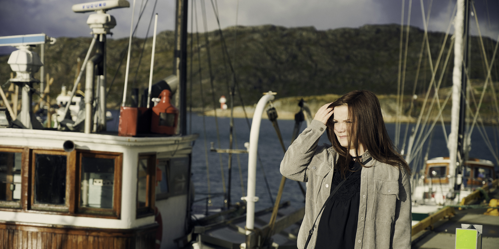 En ung kvinne med langt rødbrunt hår står i et havneområde omgitt av fiskebåter