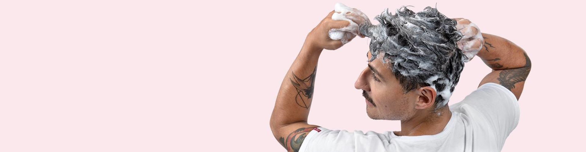 En ung mann med tatoveringer på armene vasker håret sitt med sjampo