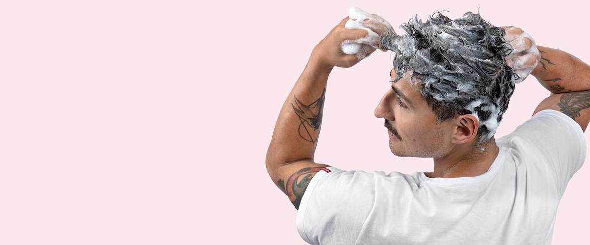 En ung mann med tatoveringer på armene vasker håret sitt med sjampo