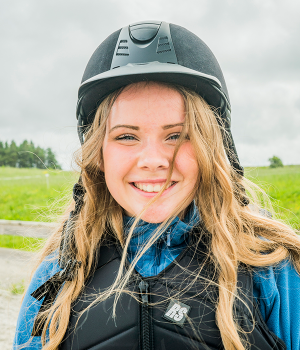 En ung jente med ridehjelm og langt blondt hår smiler mot kamera