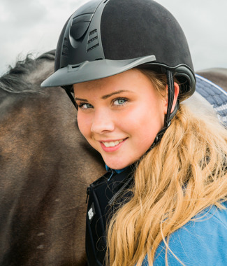 Tiril med ridehjelm og blond parykk står foran en hest